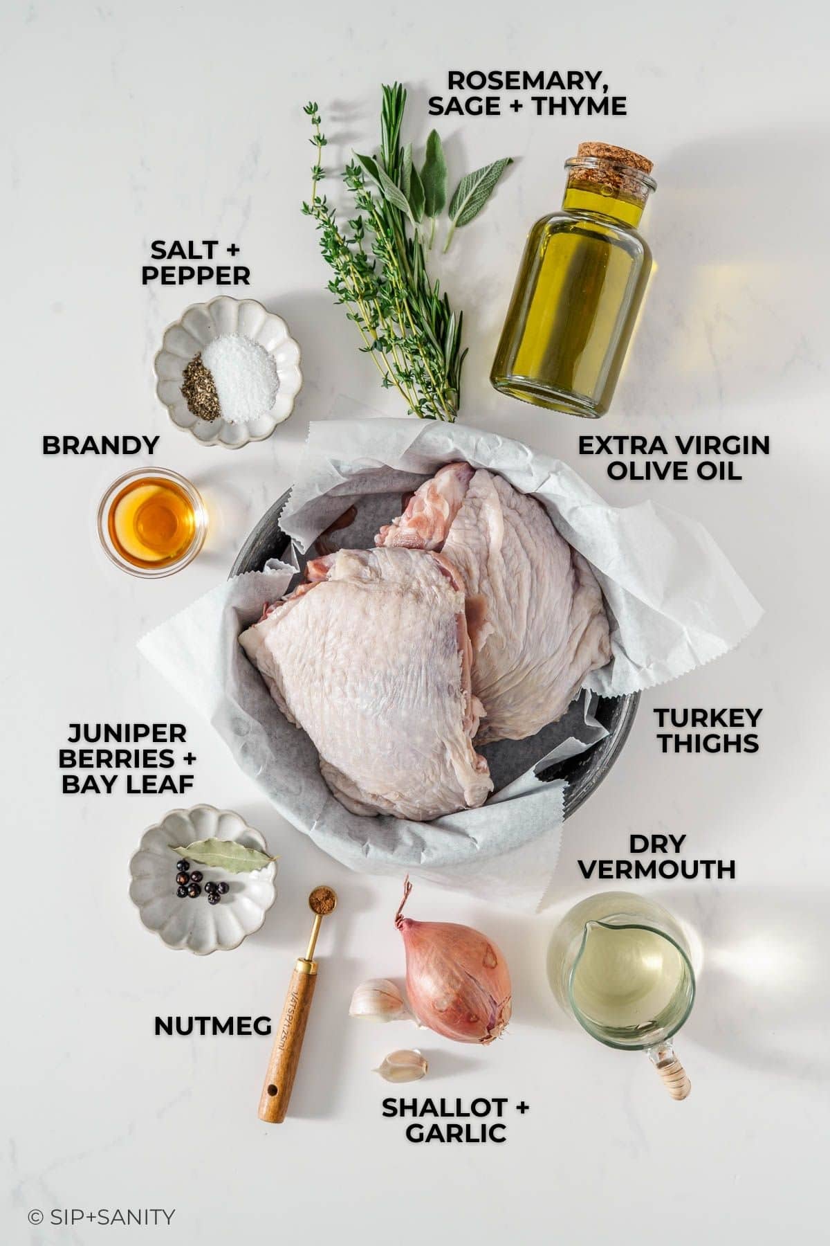 Overhead view of ingredients for cooking turkey: raw turkey thighs in a bowl, extra virgin olive oil, rosemary, sage, thyme, salt, pepper, brandy, juniper berries, bay leaf, nutmeg, shallot, garlic, and dry vermouth.