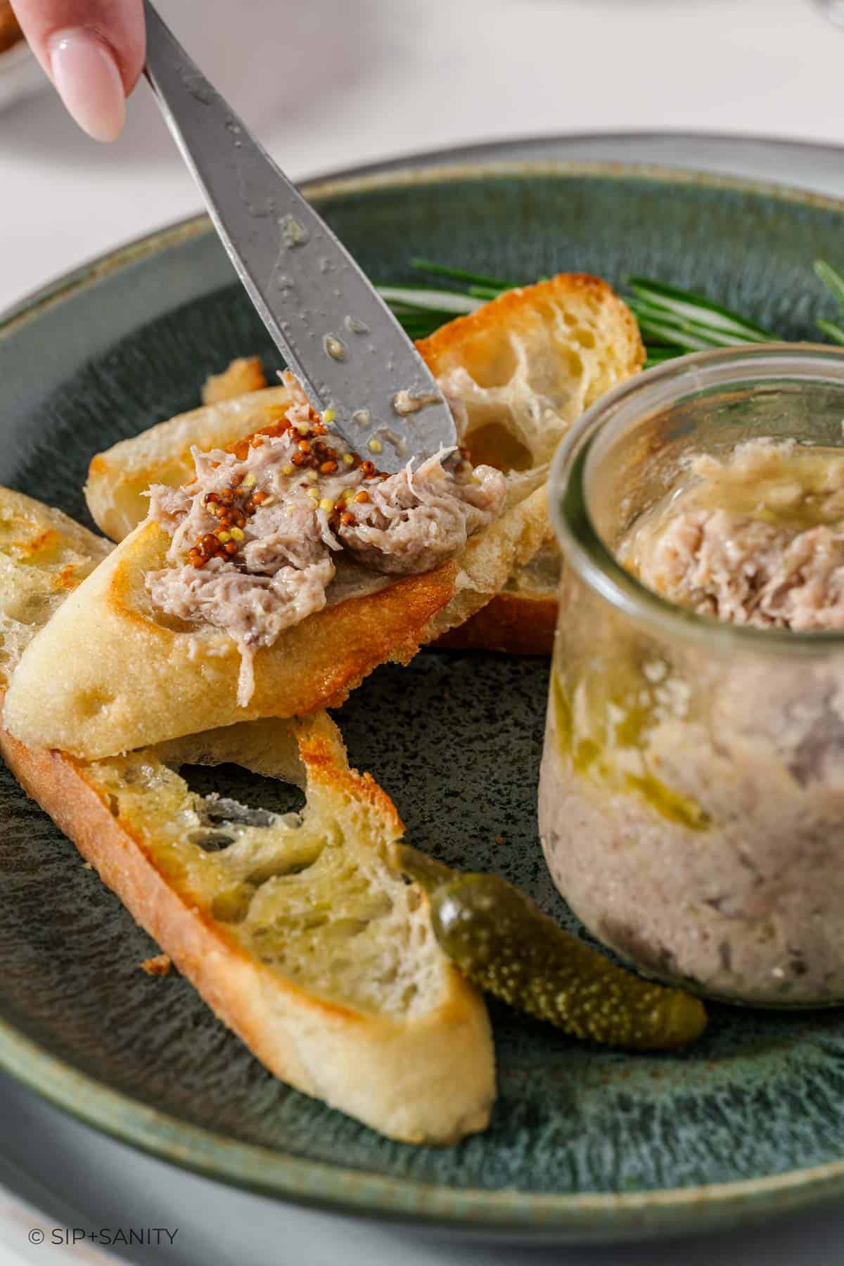 A knife spreads turkey rillettes and whole grain mustard onto toasted baguette slices, with a jar of rillettes and a cornichon on a green ceramic plate.