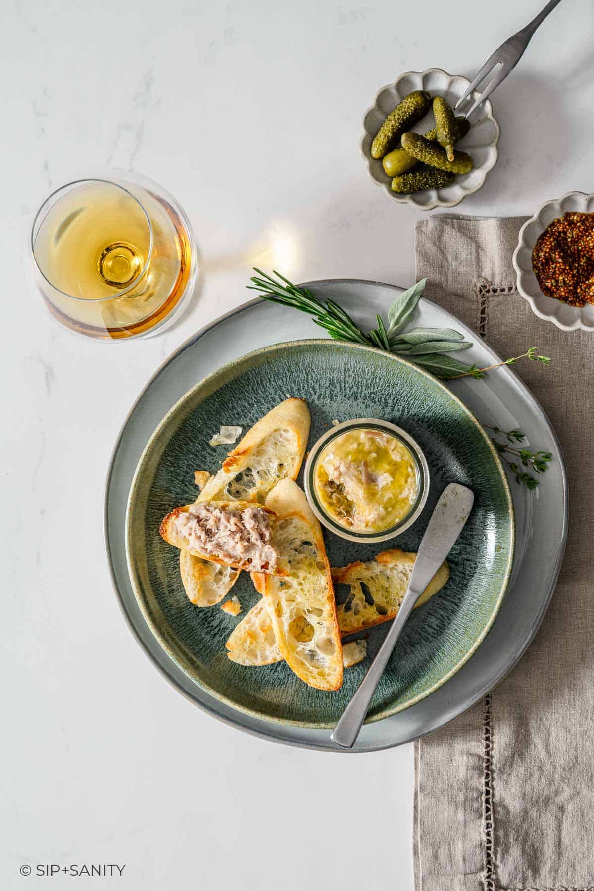 A plate with toasted baguette slices, a jar of rillettes with a knife, garnished with herbs.