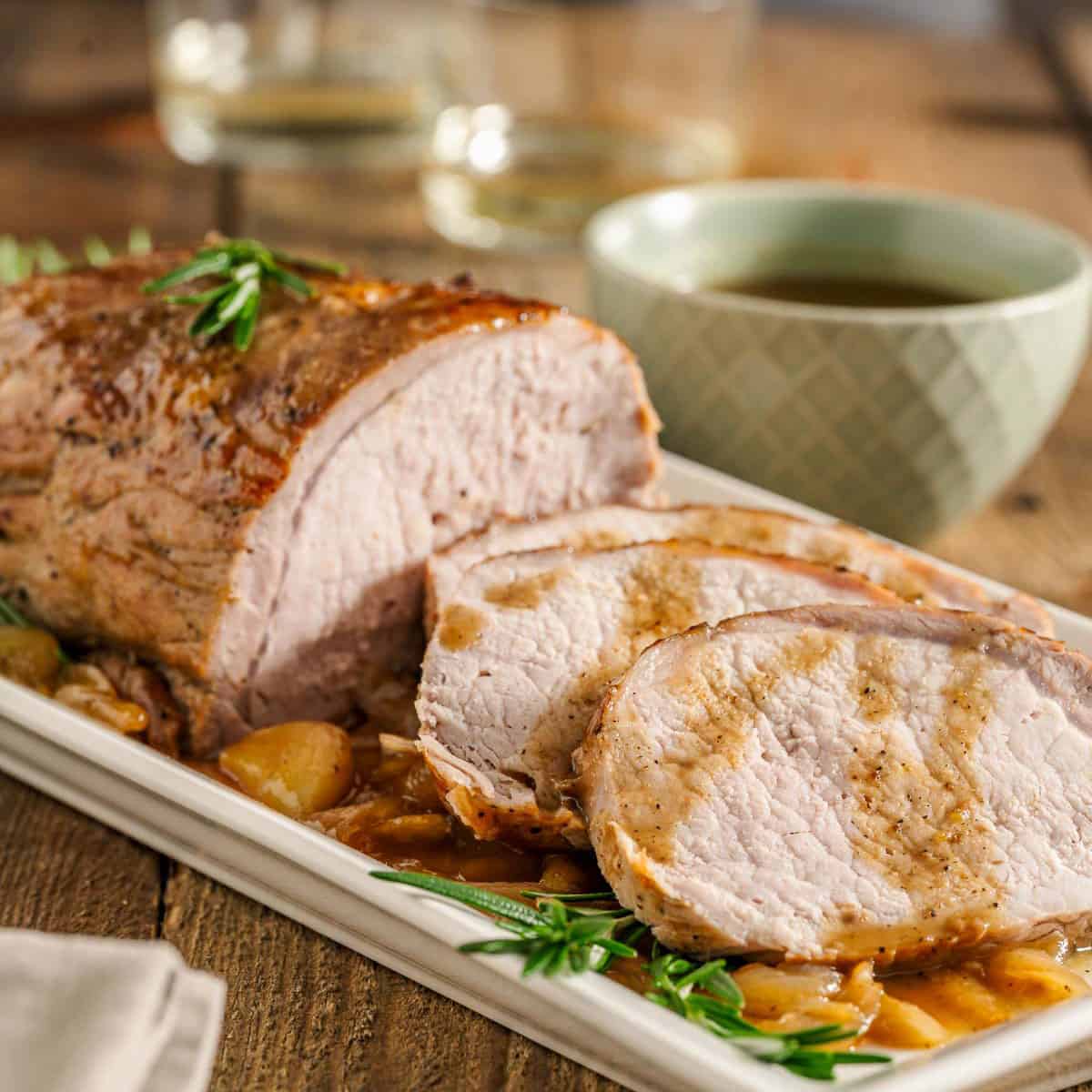 A platter with roasted pork loin slices on a bed of onions with sauce, garnished with rosemary.