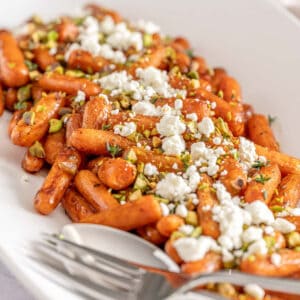 Roasted baby carrots with cheese, pistachios, and herbs on a white dish; spoon and fork rest at the edge.