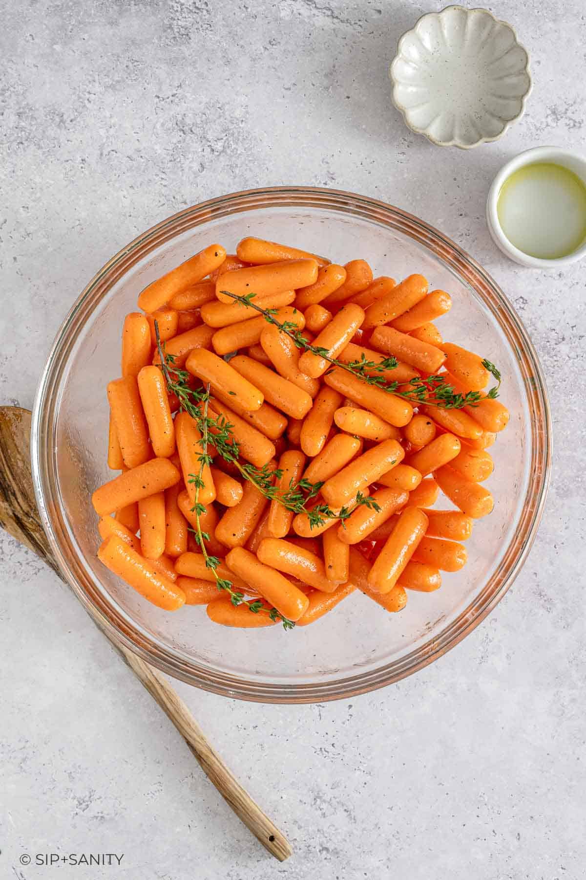 Glass bowl of seasoned baby carrots with thyme on a light counter, beside a wooden spoon and small bowls of oil and seasoning.