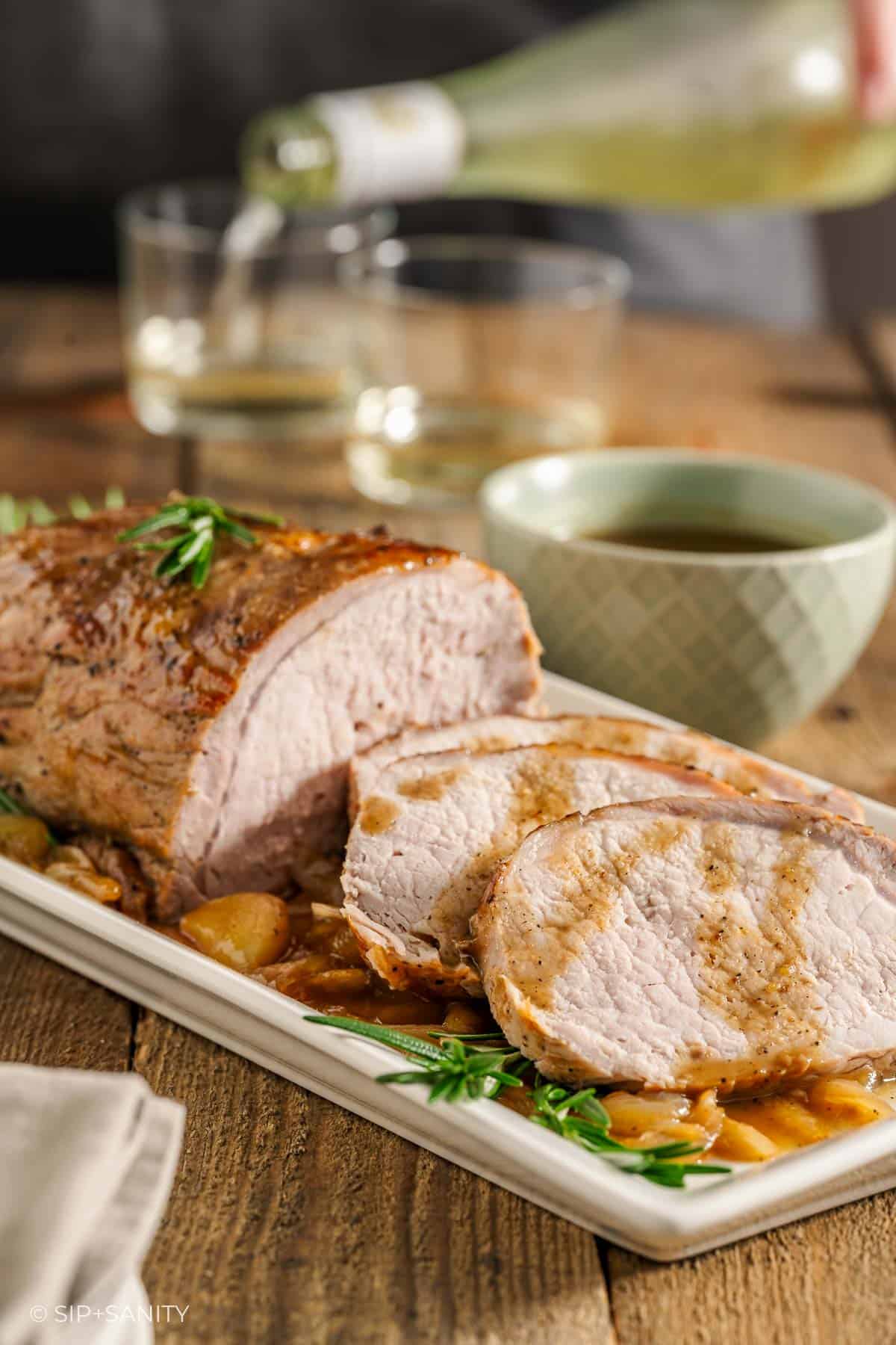 A sliced pork roast on a platter, garnished with herbs, on a table with a bowl of sauce, and someone pouring wine into glasses.