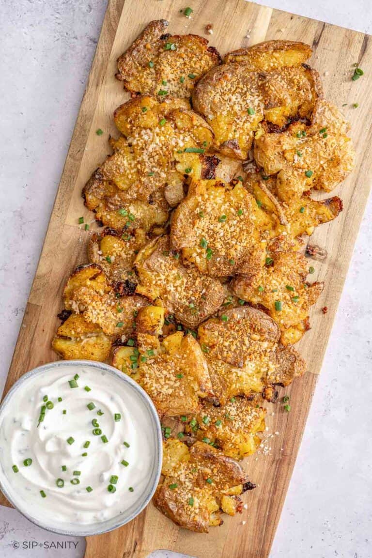 Crispy smashed potatoes with herbs and parmesan on a board, served with sour cream dip topped with chives.