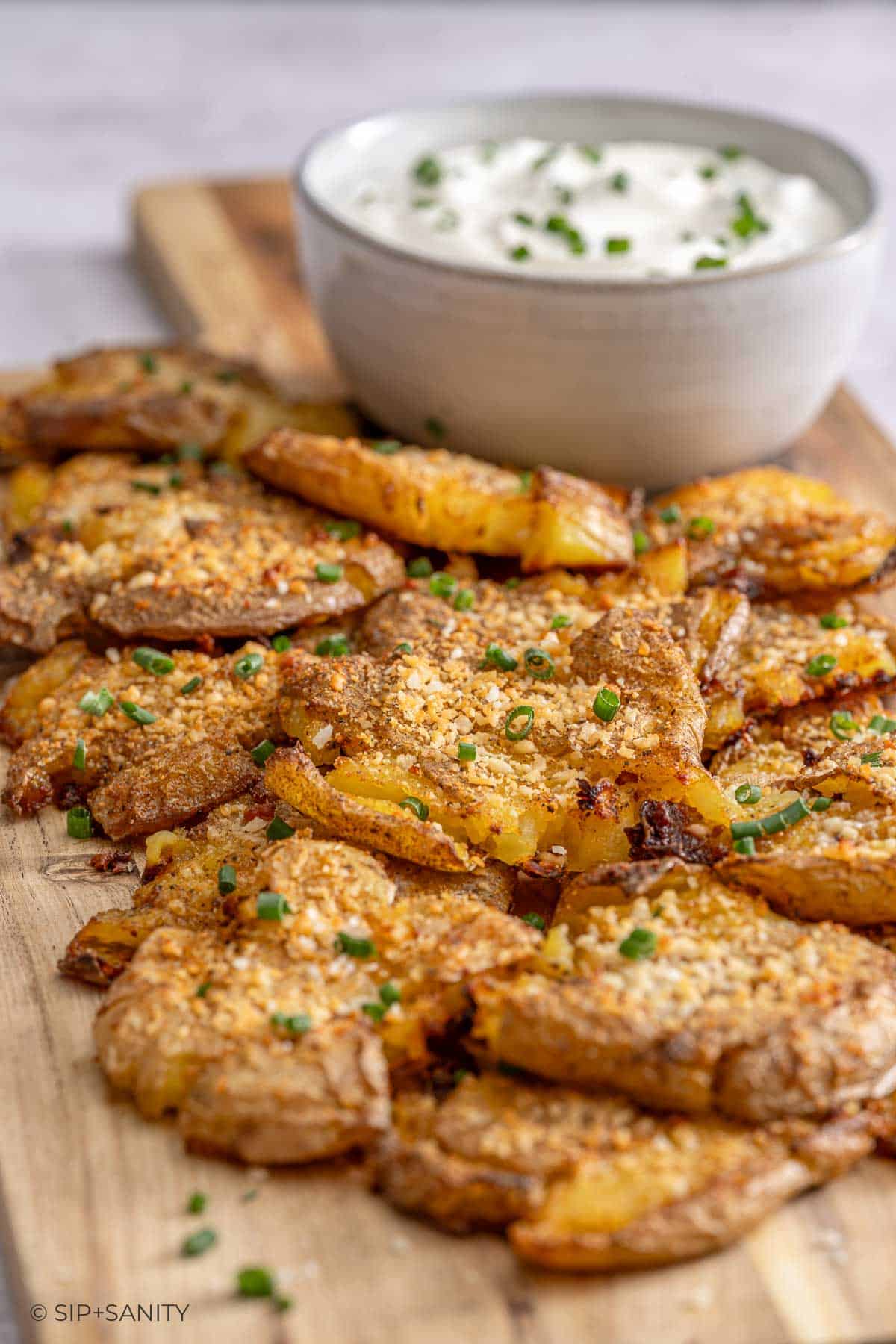 Crispy smashed potatoes with chopped chives on a wooden board, served next to a bowl of creamy dip with chives.