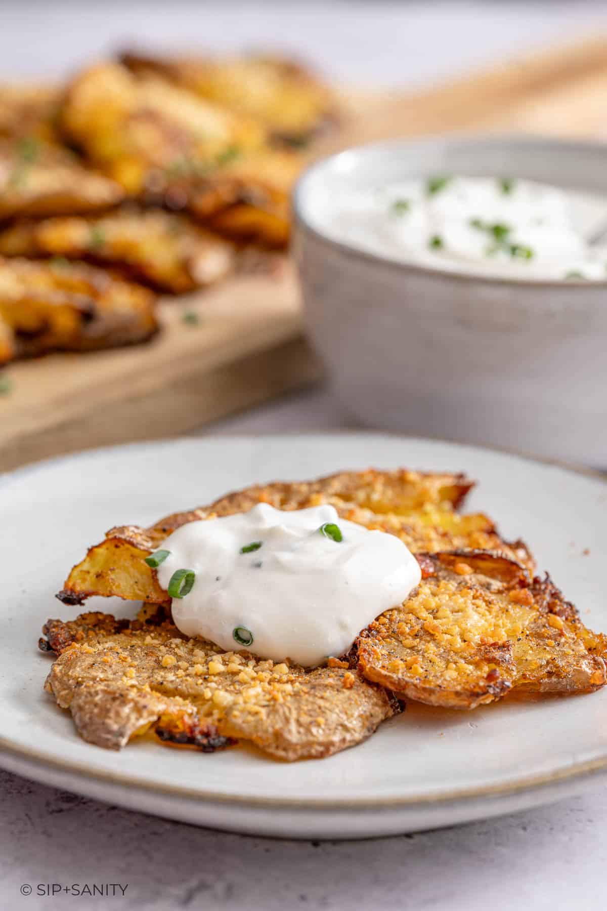 Crispy smashed potatoes with sour cream sauce and chives on a plate; more potatoes and a bowl of sauce in the background.