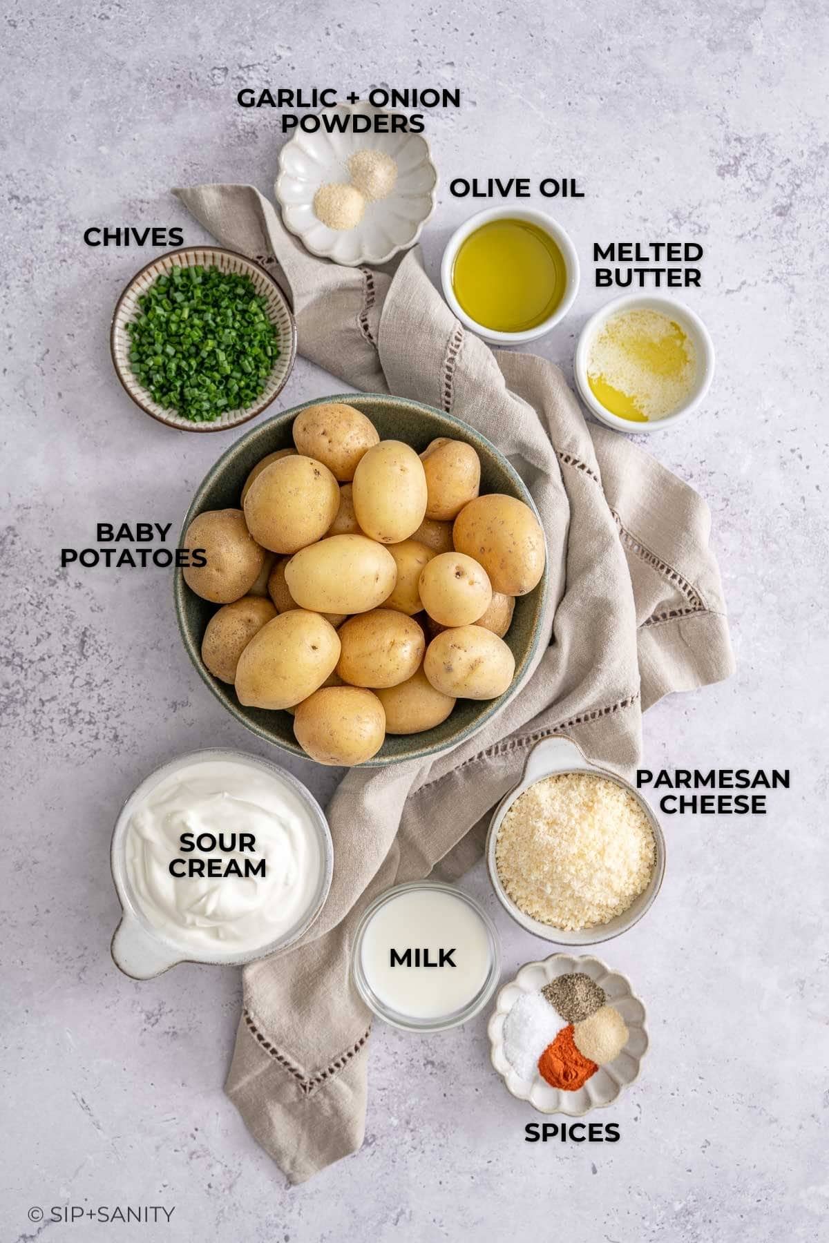 A bowl of baby potatoes with small bowls of chives, oil, butter, parmesan, sour cream, milk, and spices nearby.