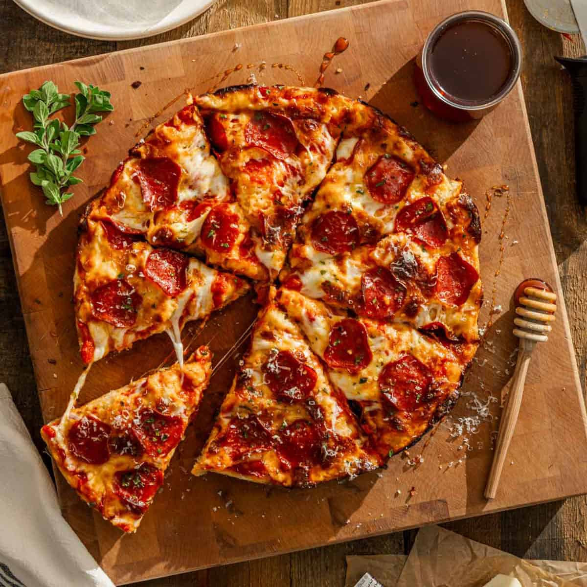 Pepperoni pizza on a board, one slice pulled away, next to a jar of honey, honey dipper, herbs, and napkin on rustic table.