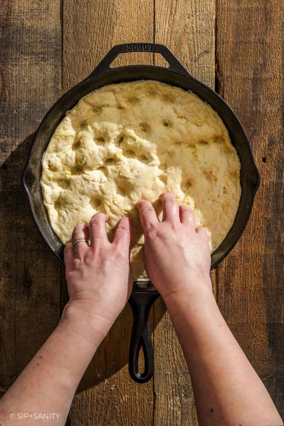 Hands docking dough into a cast iron skillet on a rustic wood surface, getting it ready to bake.