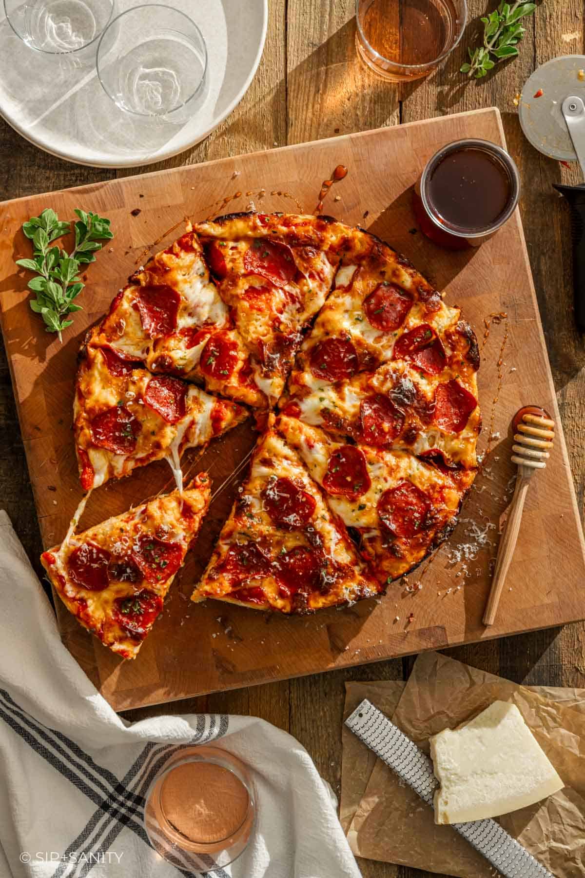 Overhead view of sliced pepperoni pizza on wooden board with herbs, drinks, cheese, grater, and utensils on rustic table.