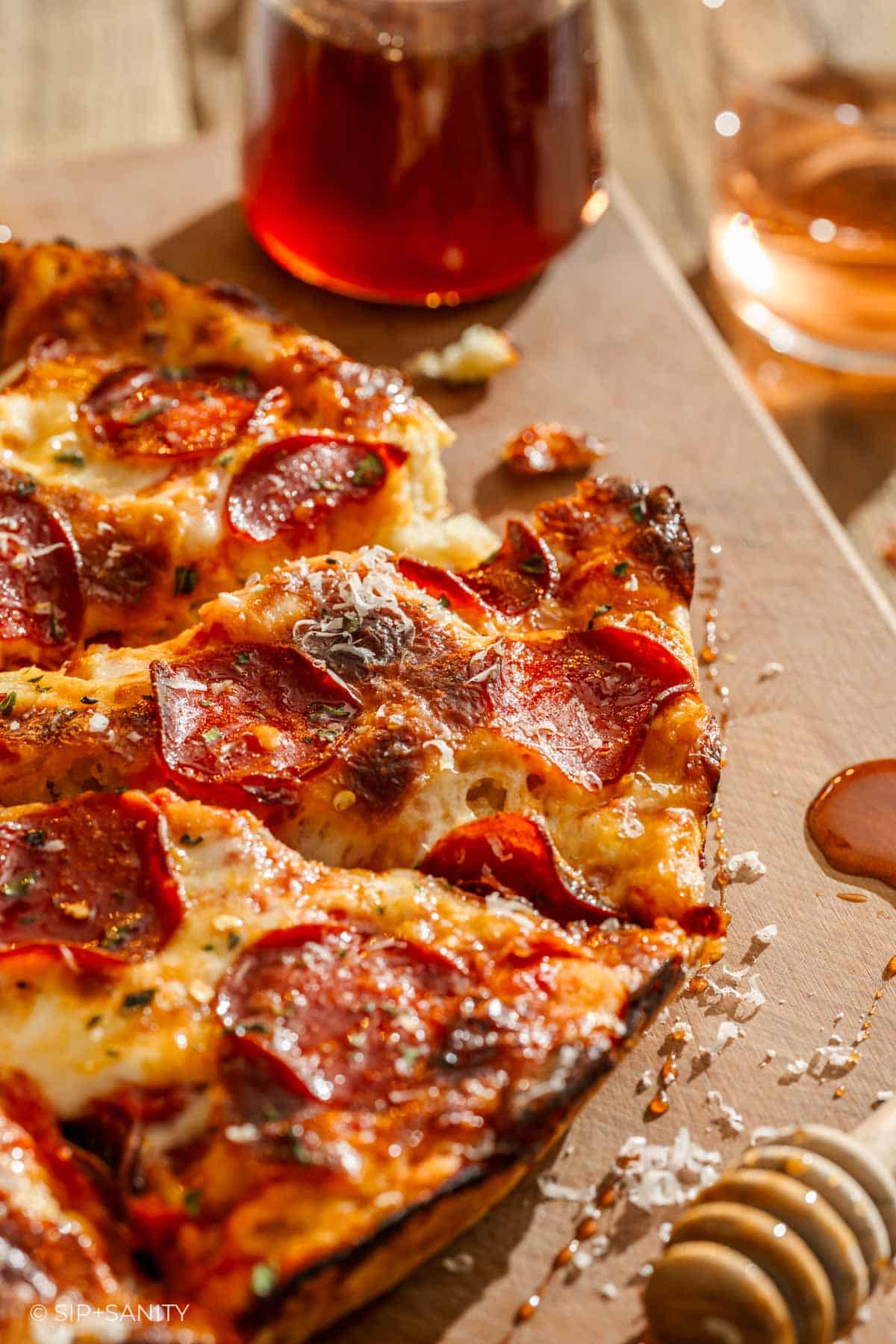 Sliced pepperoni pizza with cheese and herbs on a board; honey dipper and jar in the background.