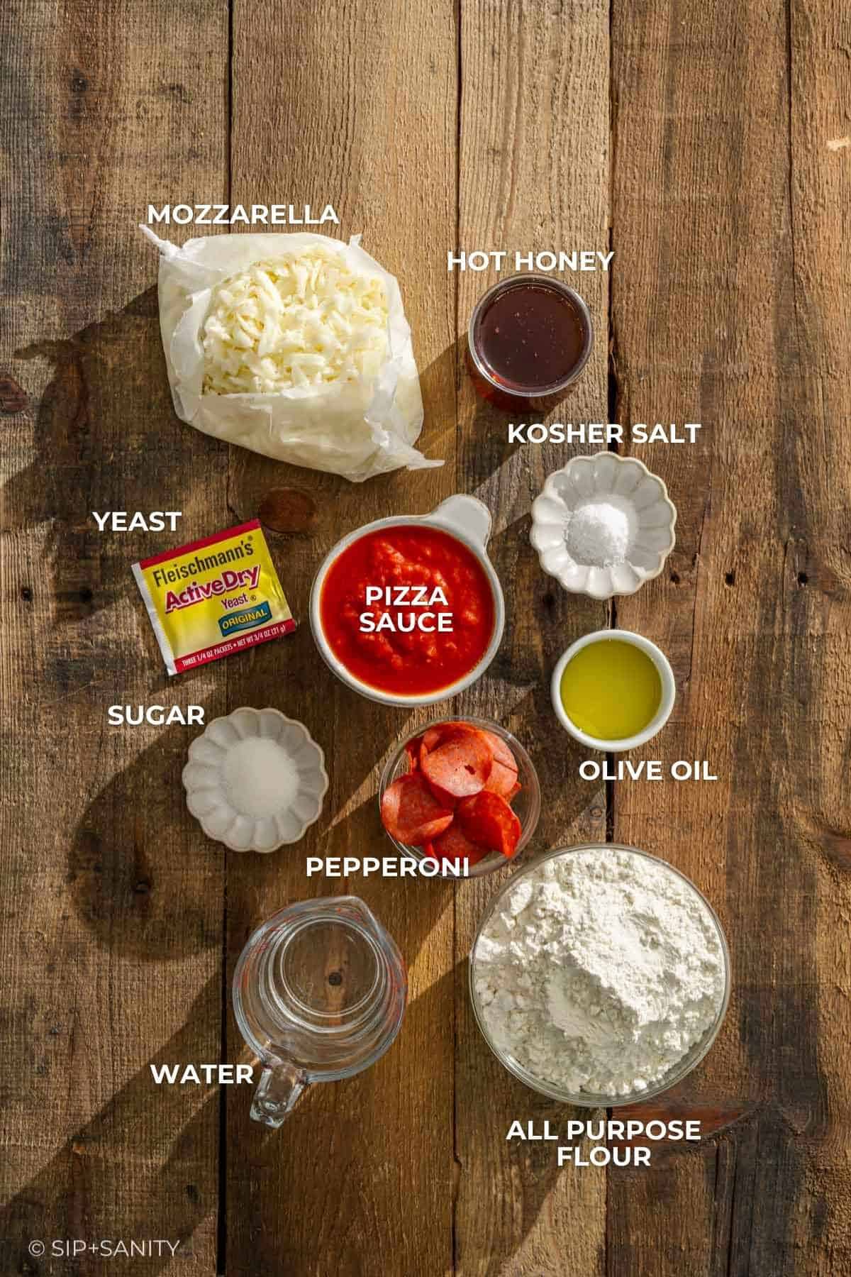 A wooden table with labeled bowls holding mozzarella, hot honey, salt, yeast, sugar, sauce, oil, pepperoni, flour, and water.