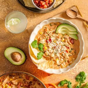 Chicken taco soup with avocado, cilantro, tortilla chips, cheese; wooden spoon, avocado half, drink, salsa nearby.