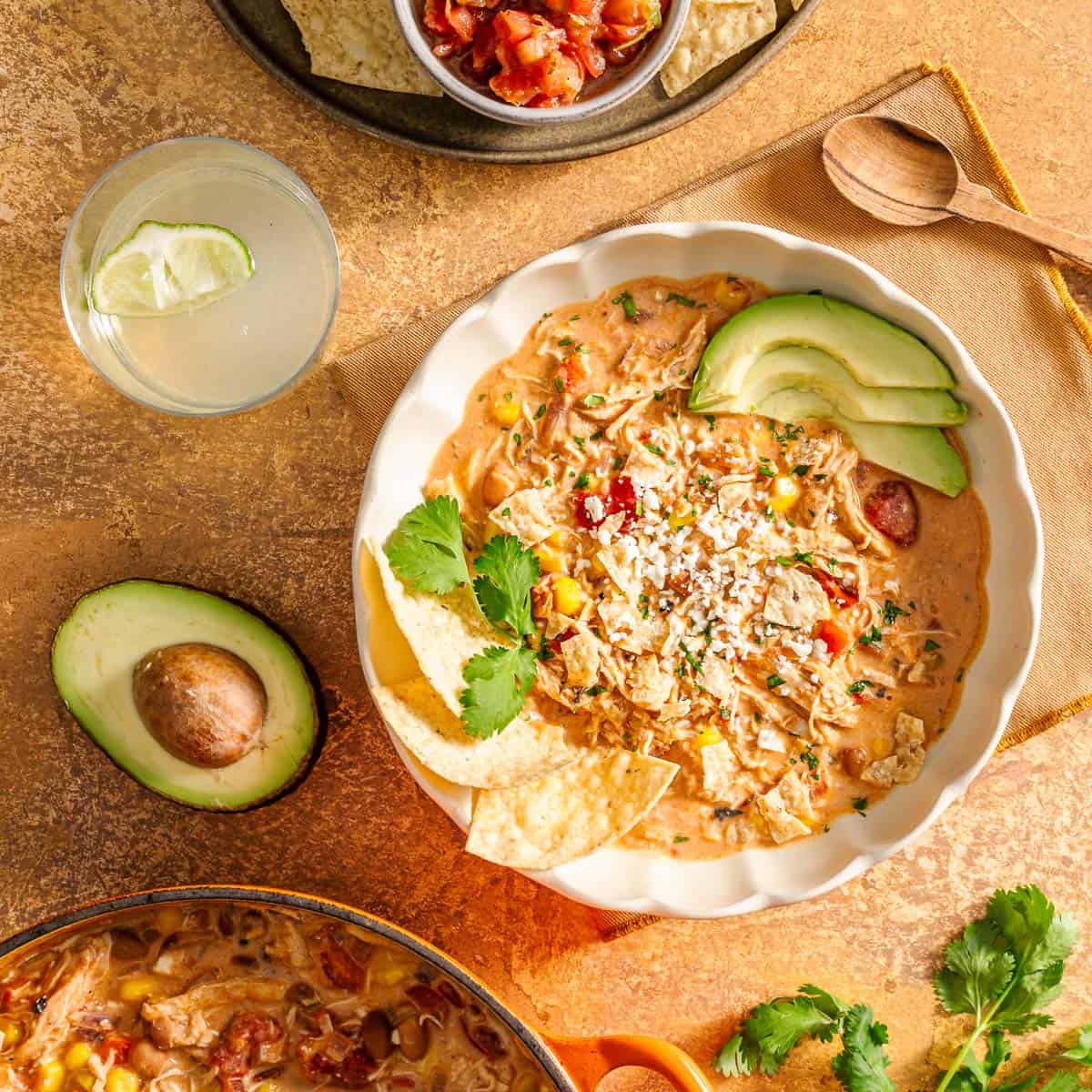 Chicken taco soup with avocado, cilantro, tortilla chips, cheese; wooden spoon, avocado half, drink, salsa nearby.
