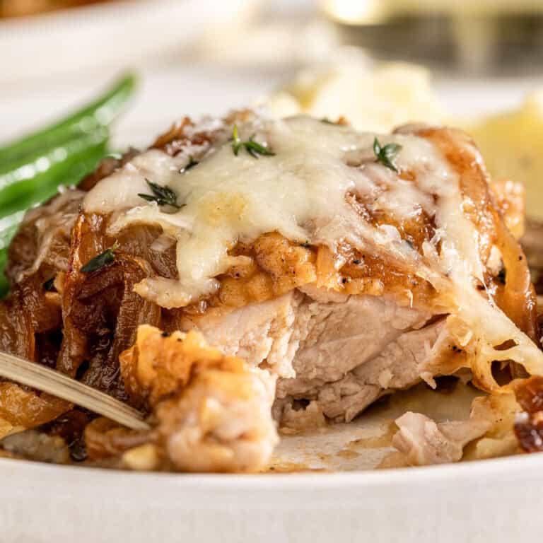 Cooked chicken thigh with melted cheese, caramelized onions, and herbs on a plate with green beans and mashed potatoes.