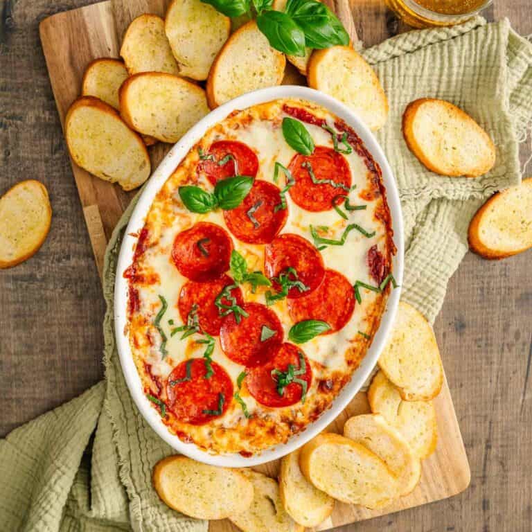 Cheesy pepperoni dip in an oval dish with basil, served with baguette slices on a wooden board and green napkin.