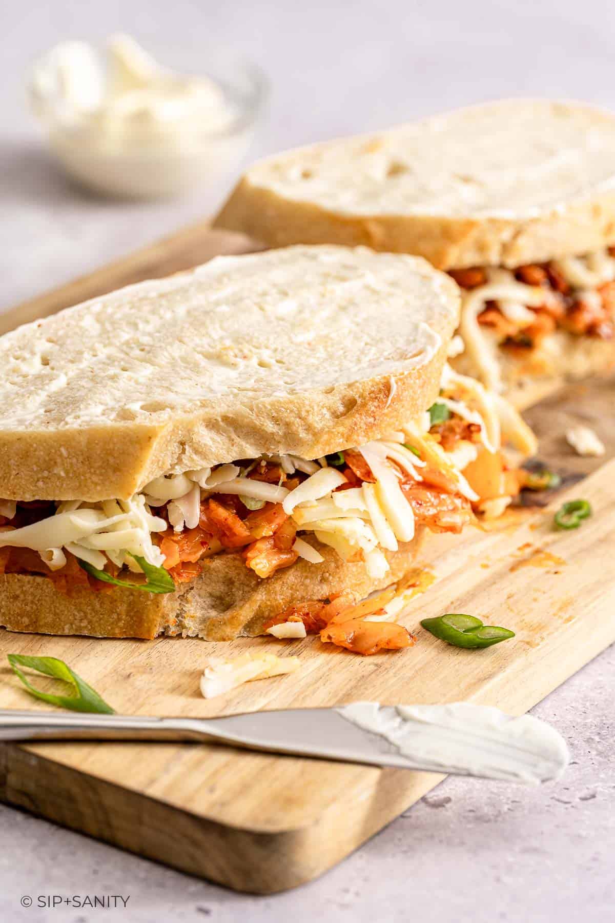 Two sandwiches with cheese and kimchi on a wooden board, with a knife and small bowl of creamy spread in the background.