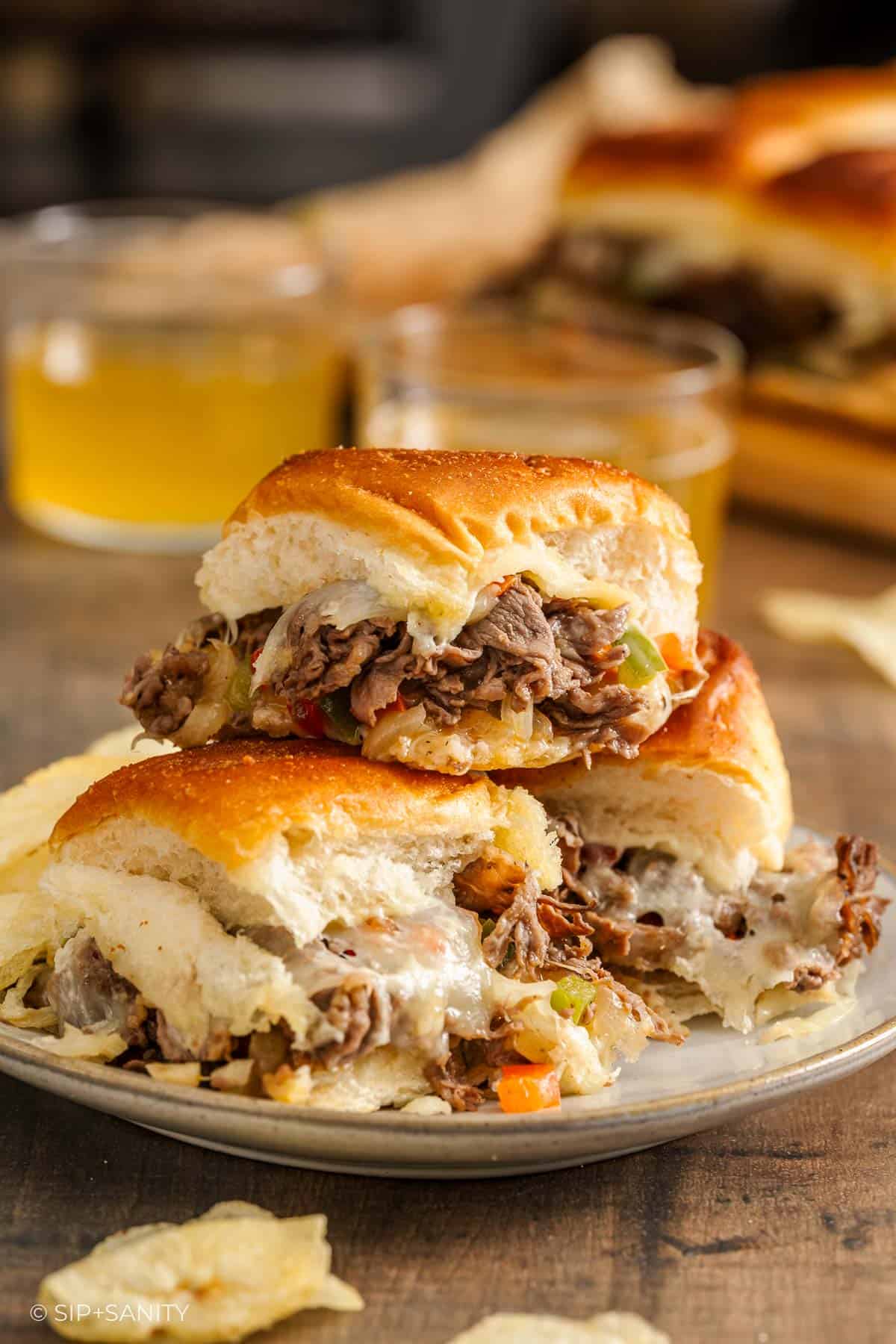Cheesesteak sliders with beef, cheese, onions, and peppers on buns, plated with drink and more sliders behind.