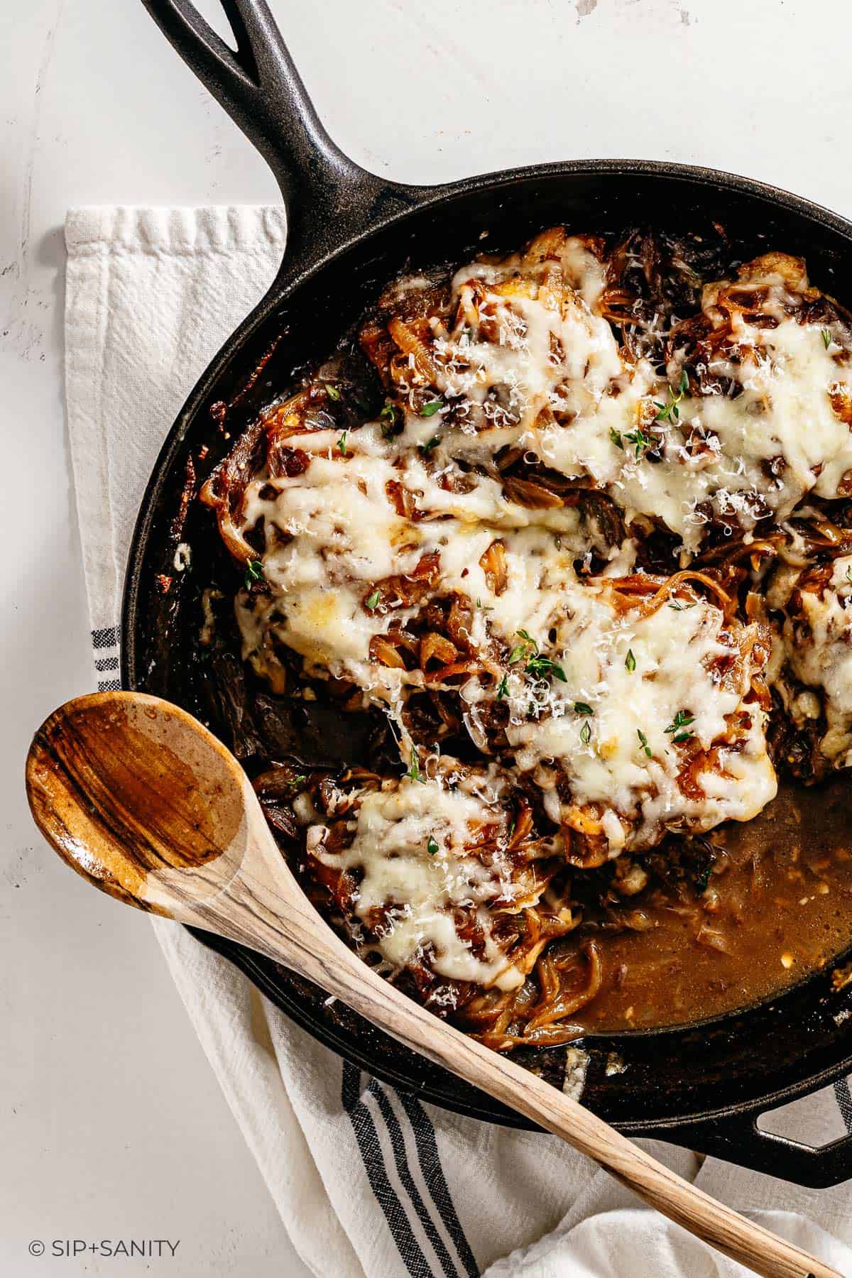 A cast-iron skillet of cheesy French onion chicken with herbs sits on a white towel, wooden spoon on the skillet’s edge.