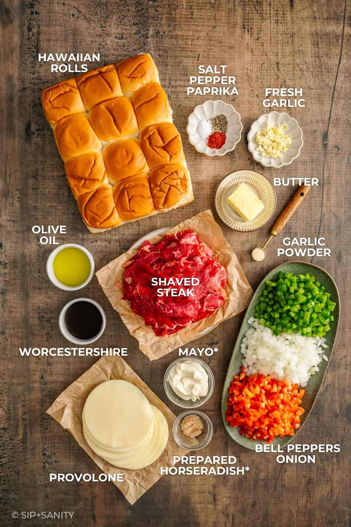 Cheesesteak slider ingredients on wood: Hawaiian rolls, shaved steak, provolone, peppers, onions, butter, oil, mayo, spices.
