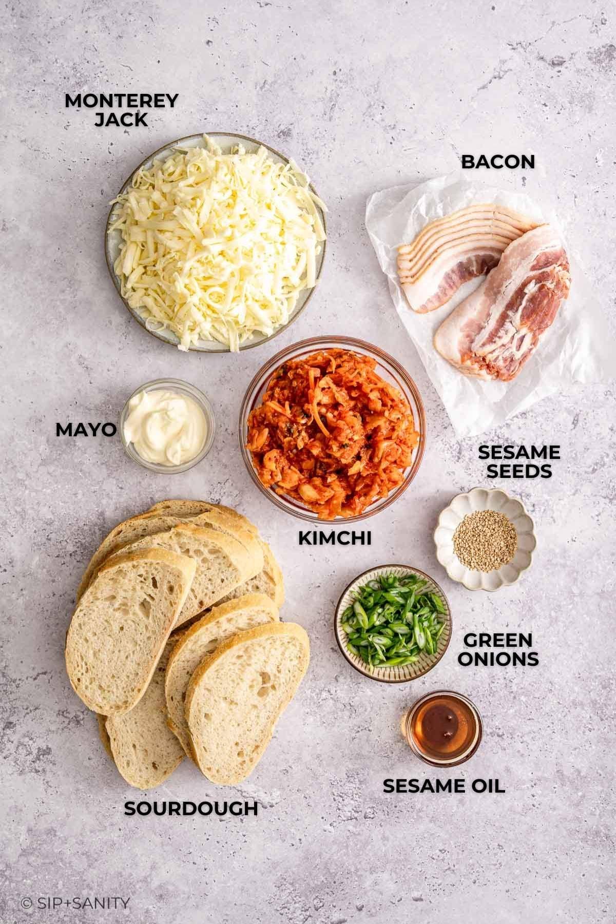 Sandwich fixings: shredded cheese, bacon, mayo, kimchi, sesame seeds, green onions, oil, sourdough on a light surface.