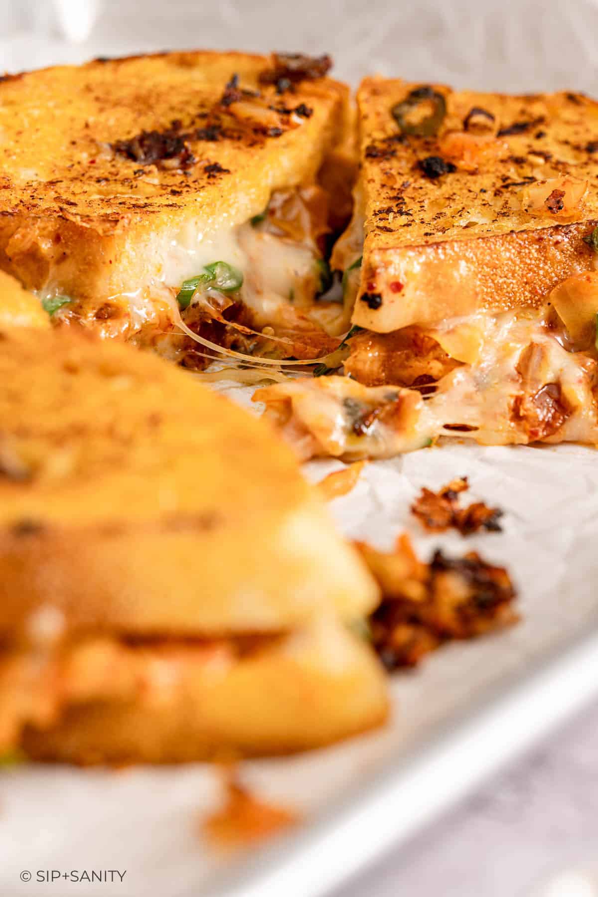 Toasted sandwich halves with melted cheese, herbs, and kimchi oozing out; crisp bread and burnt bits on white surface.