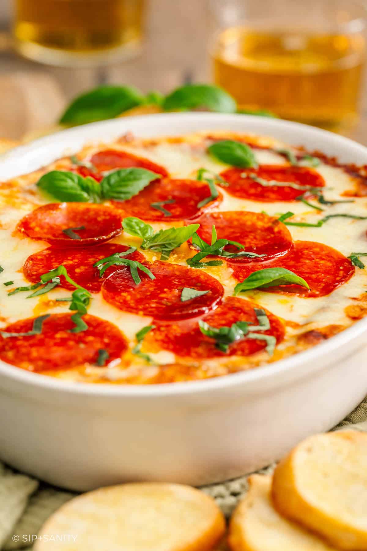 Baked pizza dip with pepperoni and basil in a white dish, crostini and beer visible in the background.