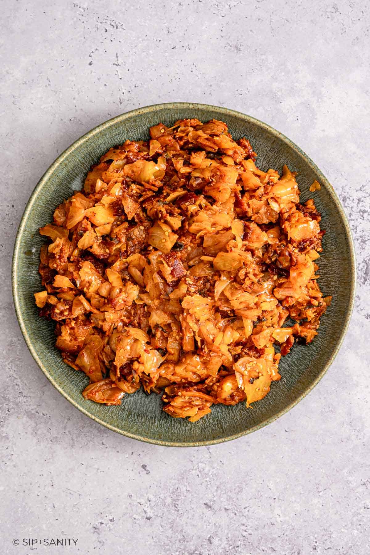 Cooked kimchi with a reddish-orange hue in a green bowl on a light gray textured surface.