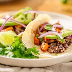 Two soft tacos with black beans, sweet potatoes, red onion, and avocado, topped with cilantro and lime on a white plate.