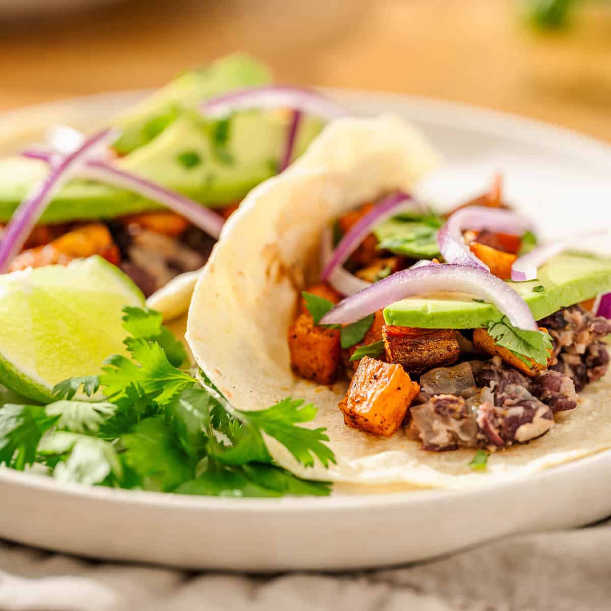 Two soft tacos with black beans, sweet potatoes, red onion, and avocado, topped with cilantro and lime on a white plate.