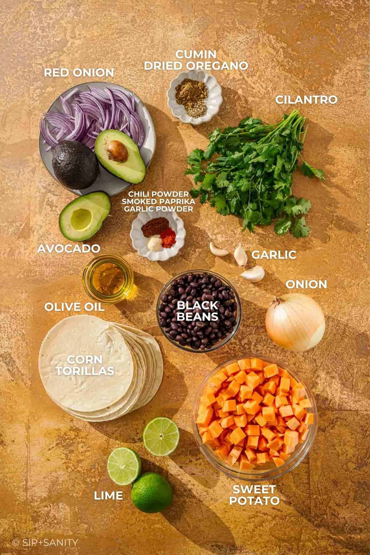Assorted labeled ingredients on brown surface: onion, spices, herbs, avocado, beans, tortillas, lime, and sweet potato.