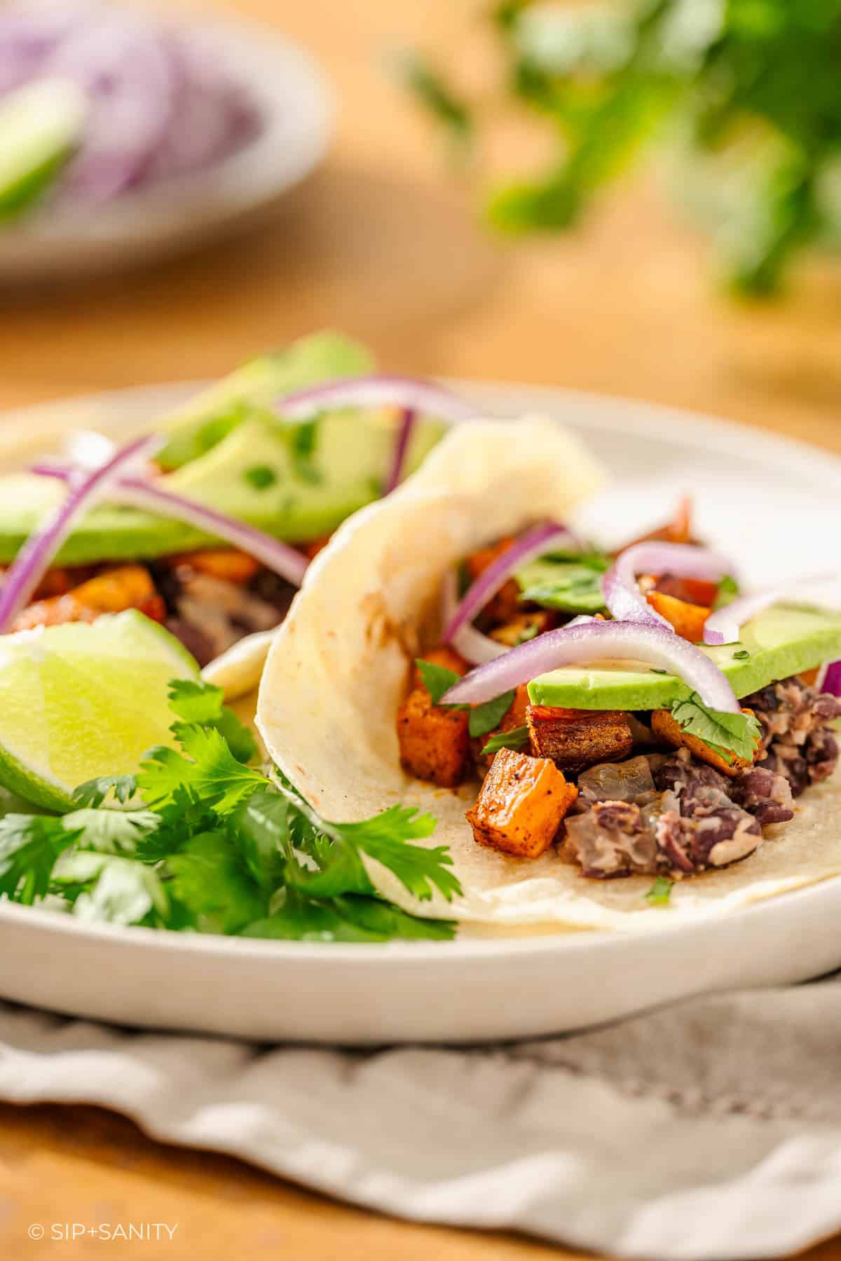 Two soft tacos with black beans, sweet potatoes, avocado, red onion, cilantro, and lime on a white plate.