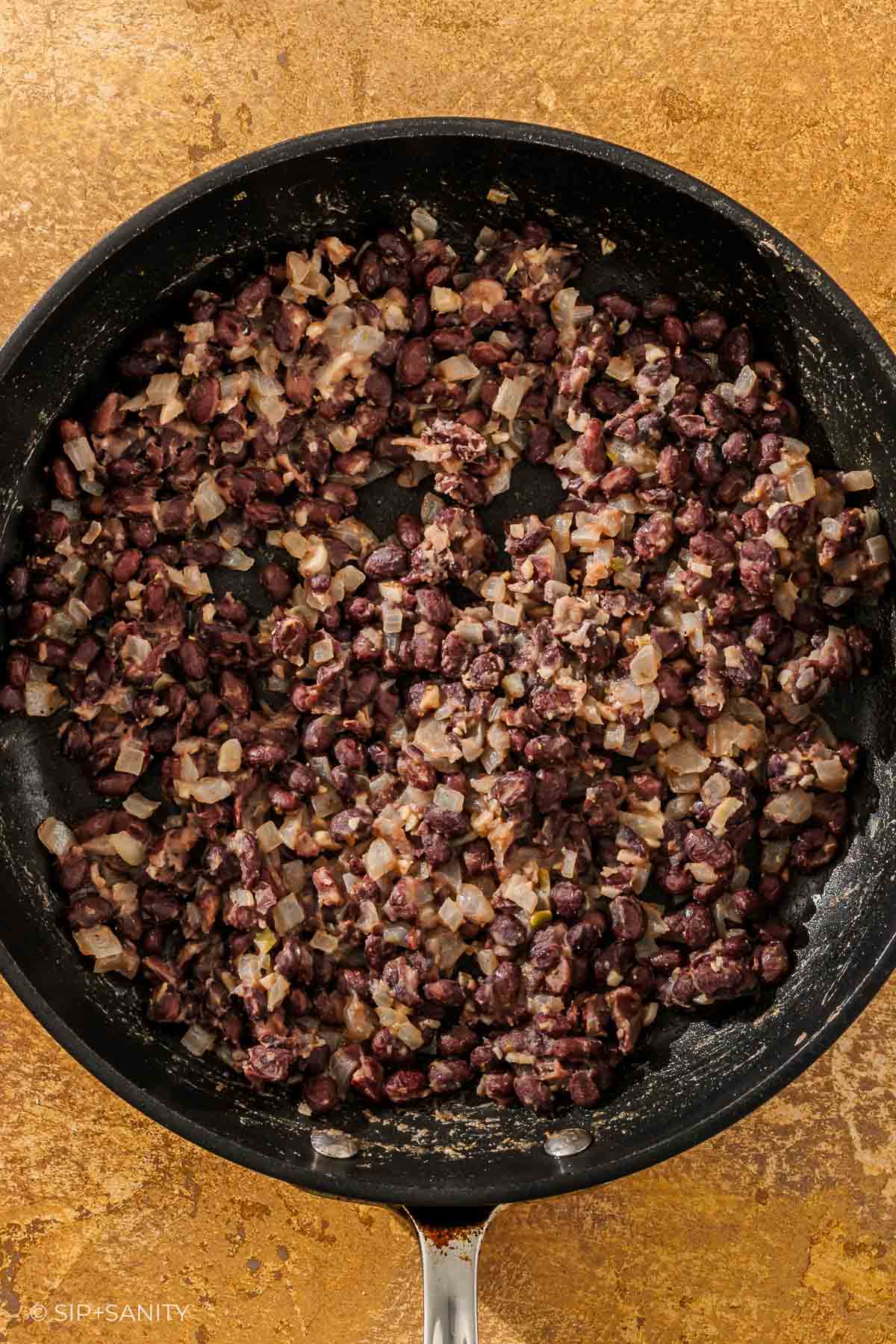 Skillet with sautéed black beans, diced onions, and seasonings viewed from above on a brown textured surface.