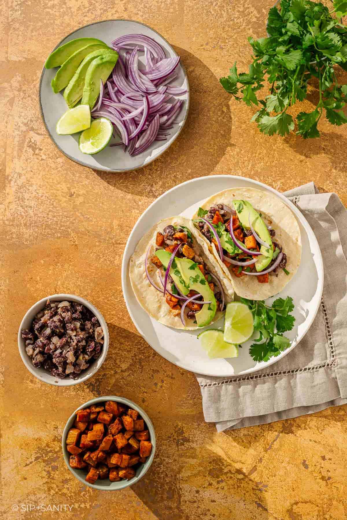 Two veggie tacos with avocado, red onion, and cilantro on a plate with lime; bowls of veggies and beans surround them.