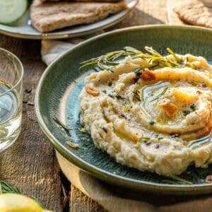 Creamy butter bean dip on a green plate, topped with oil, herbs, toasted garlic, beside wine and flatbread on a wooden table.