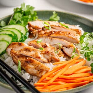 Vietnamese lemongrass chicken on white rice with green onions, cucumber, carrots, cilantro on a green plate and black chopsticks.
