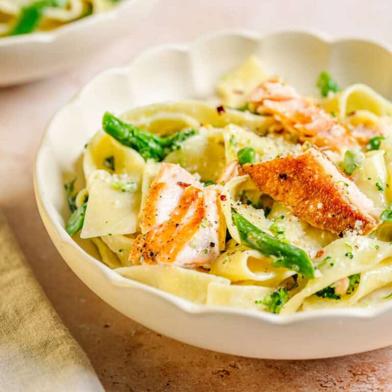 Creamy pasta with salmon, broccolini, and peas, topped with cheese in a white scalloped bowl on a light surface.