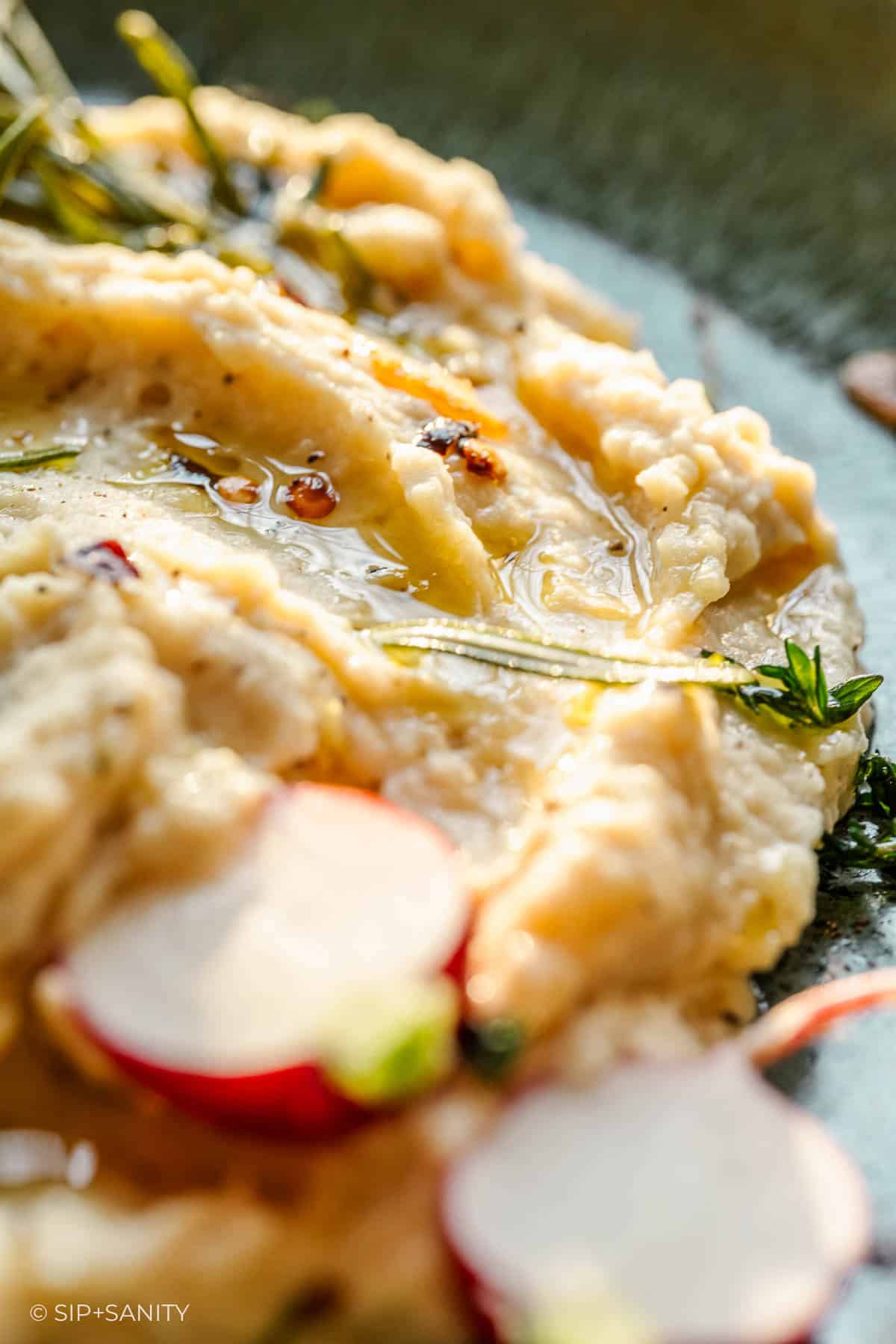 Creamy butter bean dip with olive oil, fresh herbs, and radish slices on a plate; dip texture and toppings clearly visible.