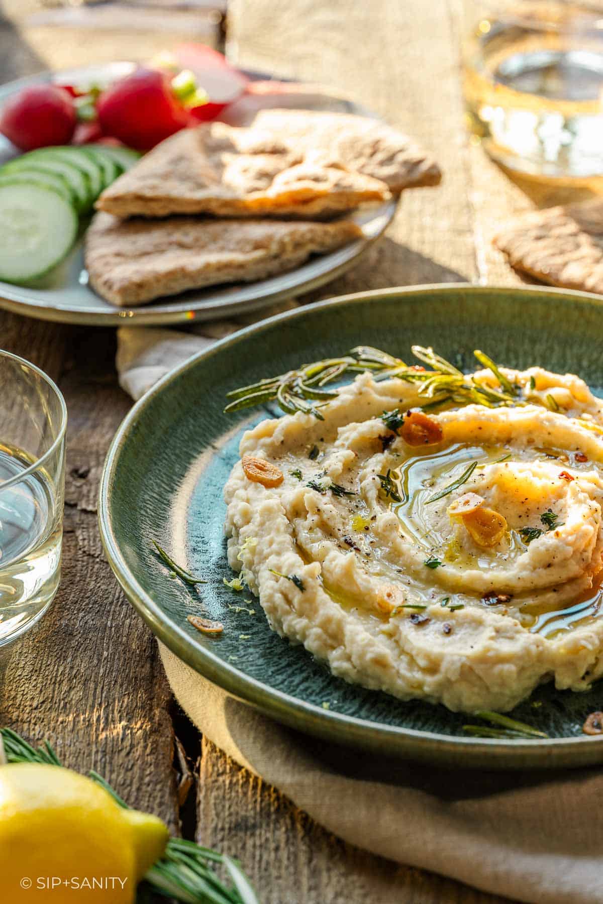 A green plate of butter bean dip with olive oil, herbs, garlic sits on wood table near veggies, pita, lemon, wine in sunlight.