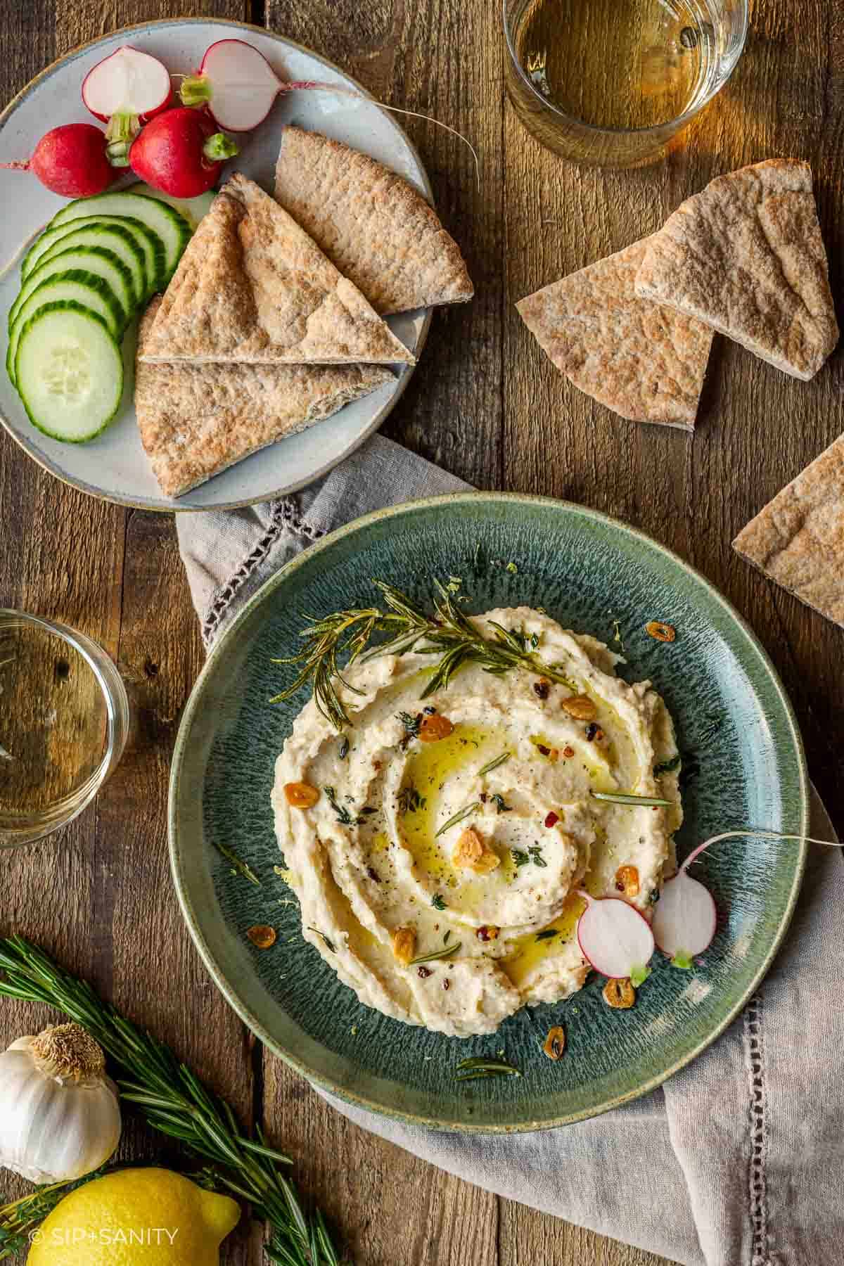 White bean dip with herbs and oil on green plate, served with pita, cucumber, radishes; wine glasses in background.