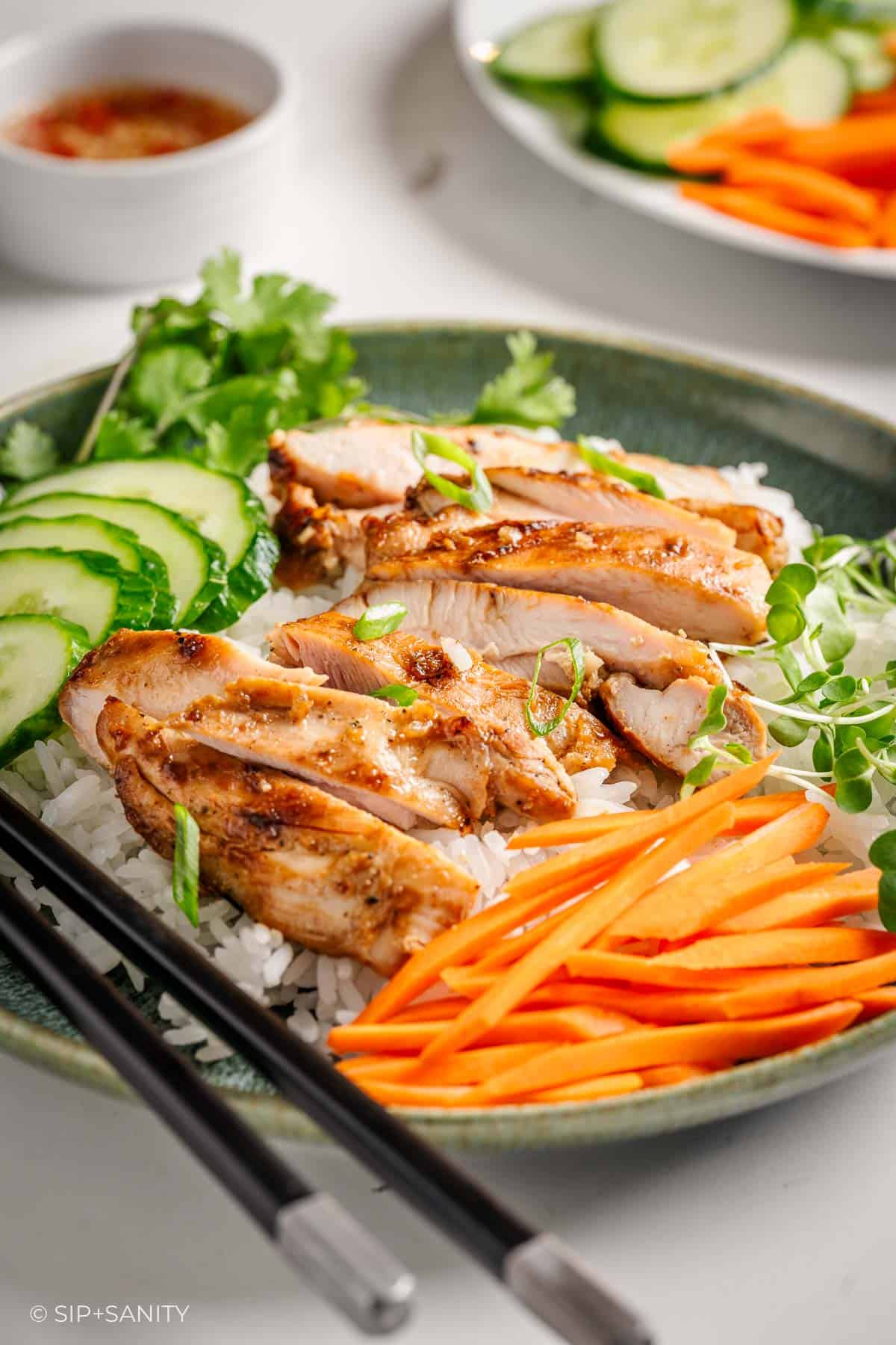 Green bowl of lemongrass chicken on rice with cucumber, carrots, cilantro, green onions, chopsticks, and dipping sauce nearby.