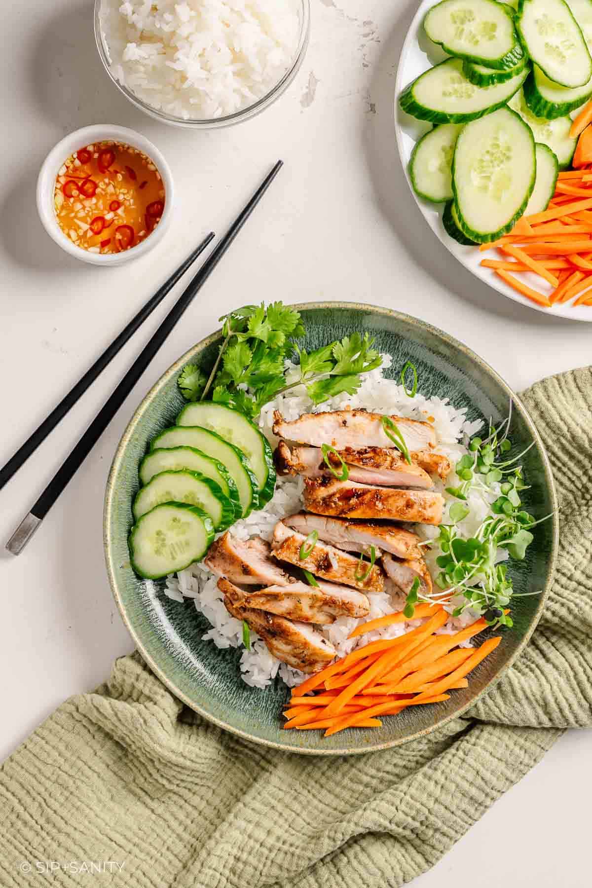 Rice bowl with vietnamese lemongrass chicken, cucumber, carrot sticks, cilantro, microgreens. Chopsticks and sauce nearby on a light surface.