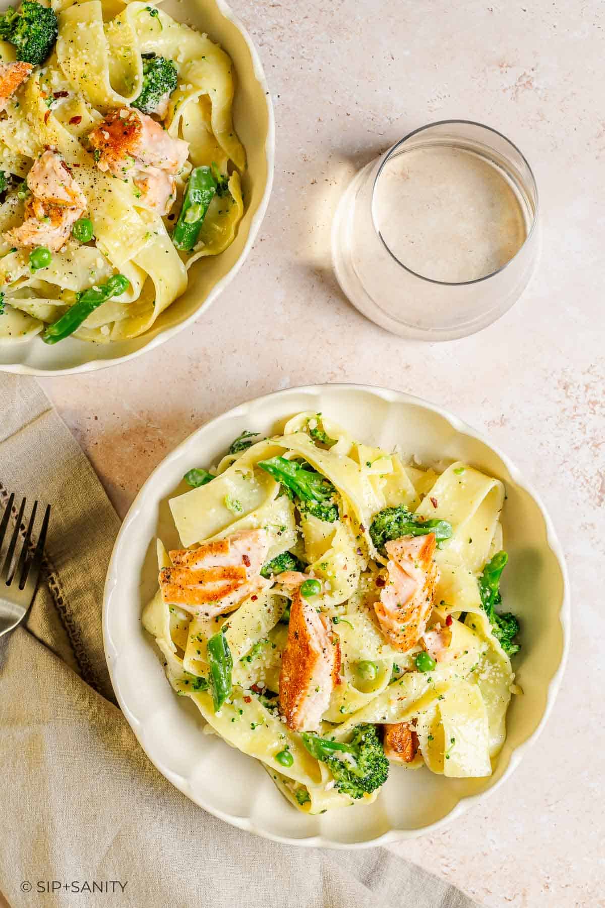 Two bowls of salmon, broccolini, and pappardelle in lemon alfredo sauce. Glass of water and fork on a napkin nearby.