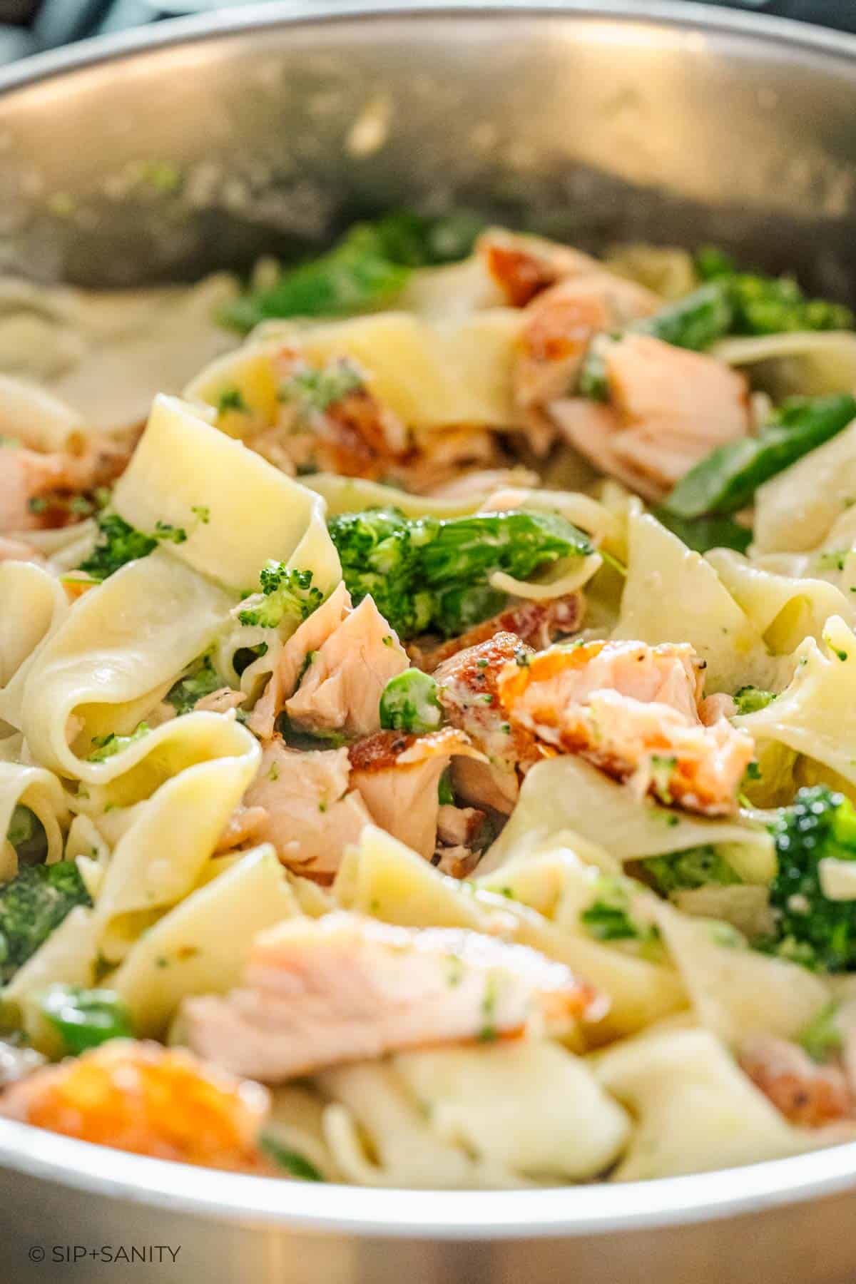 Wide noodles, salmon pieces, and broccolini mixed in lemon alfredo sauce in a pan, shown in close-up.
