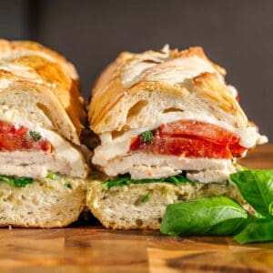 Sliced sandwich with grilled chicken, mozzarella, tomato, spinach & pesto on crusty bread. Basil beside on wooden surface.