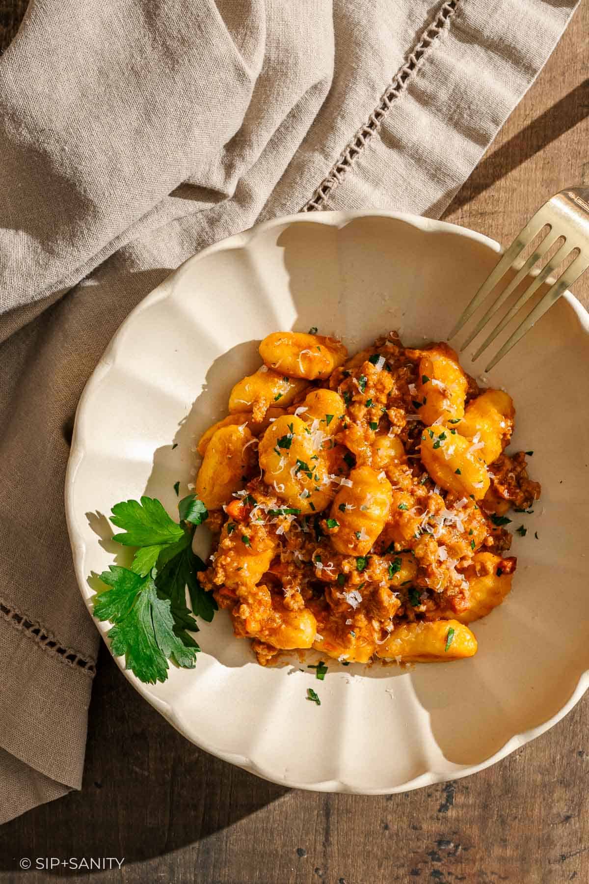 Gnocchi in bolognese with bechamel sauce with grated cheese and parsley on a white bowl, fork and linen napkin beside.