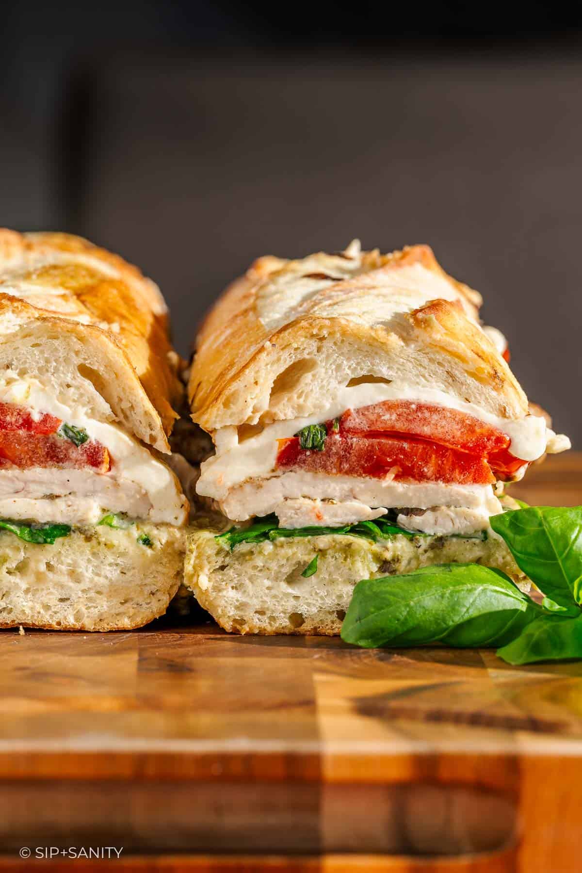 Sliced sandwich with chicken, burrata, tomato, and basil on a wooden board, with a basil sprig beside it.
