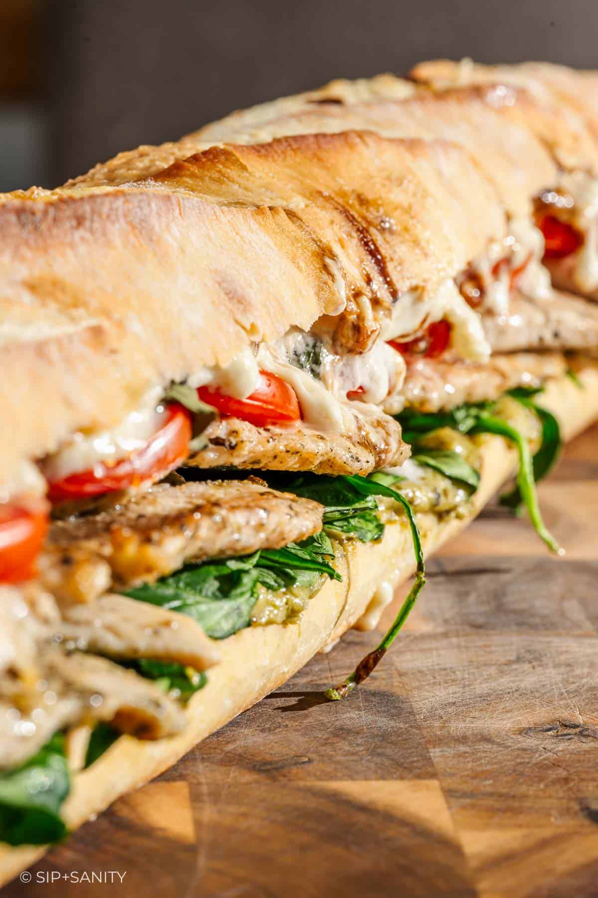 A long baguette sandwich stuffed with grilled chicken, tomato, basil pesto, greens and melty mozzarella cheese on a wood board.