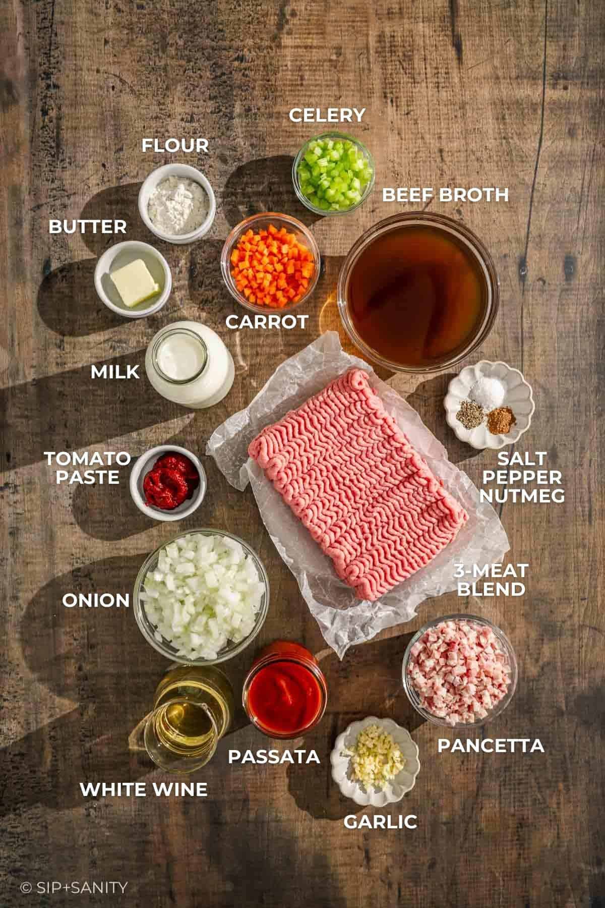 Meat sauce ingredients like ground meat, veggies, pancetta, dairy, broth, wine, tomato paste, garlic & spices on wood.