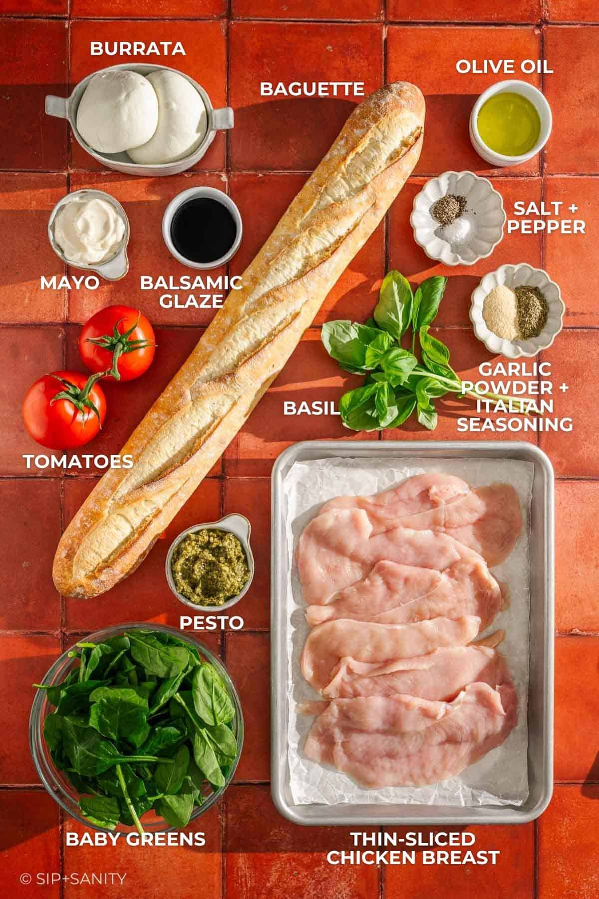 Baguette, burrata, olive oil, mayo, balsamic glaze, basil, tomato, chicken breast, pesto, greens & spices on red tiles.